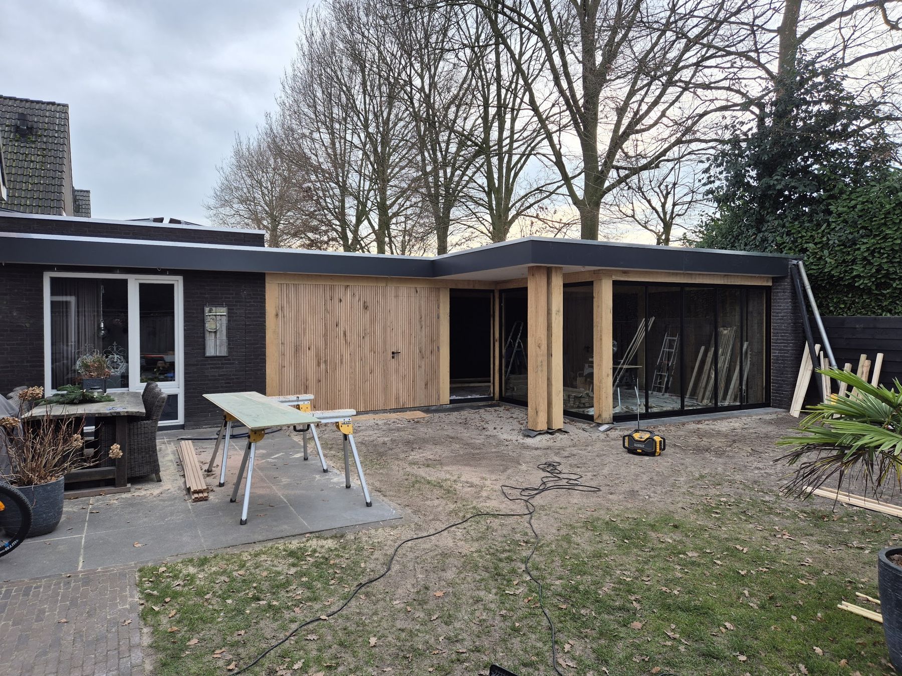Een houten aanbouw in L-vorm tijdens de bouw, met zaagtafel en gereedschap in de tuin
