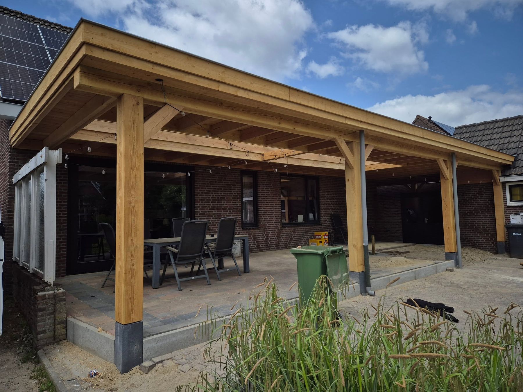 Een grote houten overkapping met zware staanders tegen een bakstenen woning, met een eettafel eronder