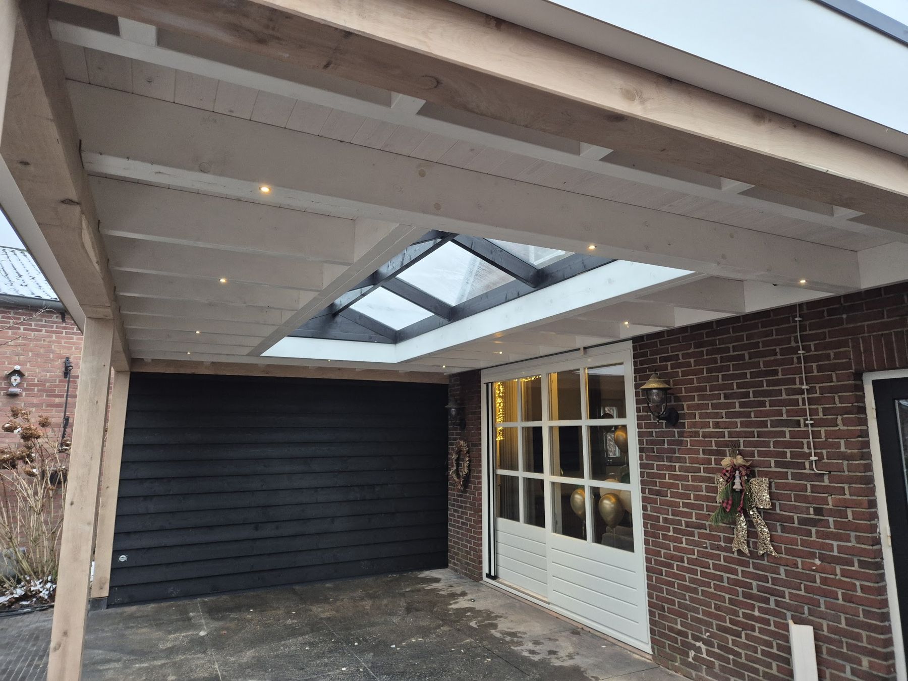 Een houten overkapping met een lichtstraat en spotjes, tussen een zwarte gevel en een bakstenen woning